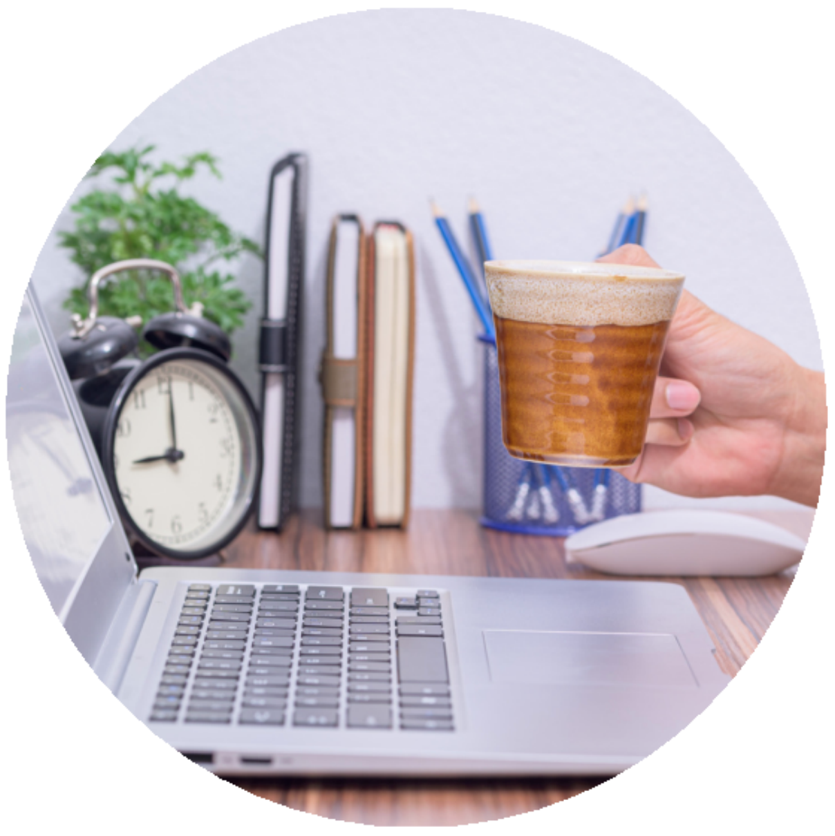 Hand holding iced coffee near a laptop on a desk with books and a clock.