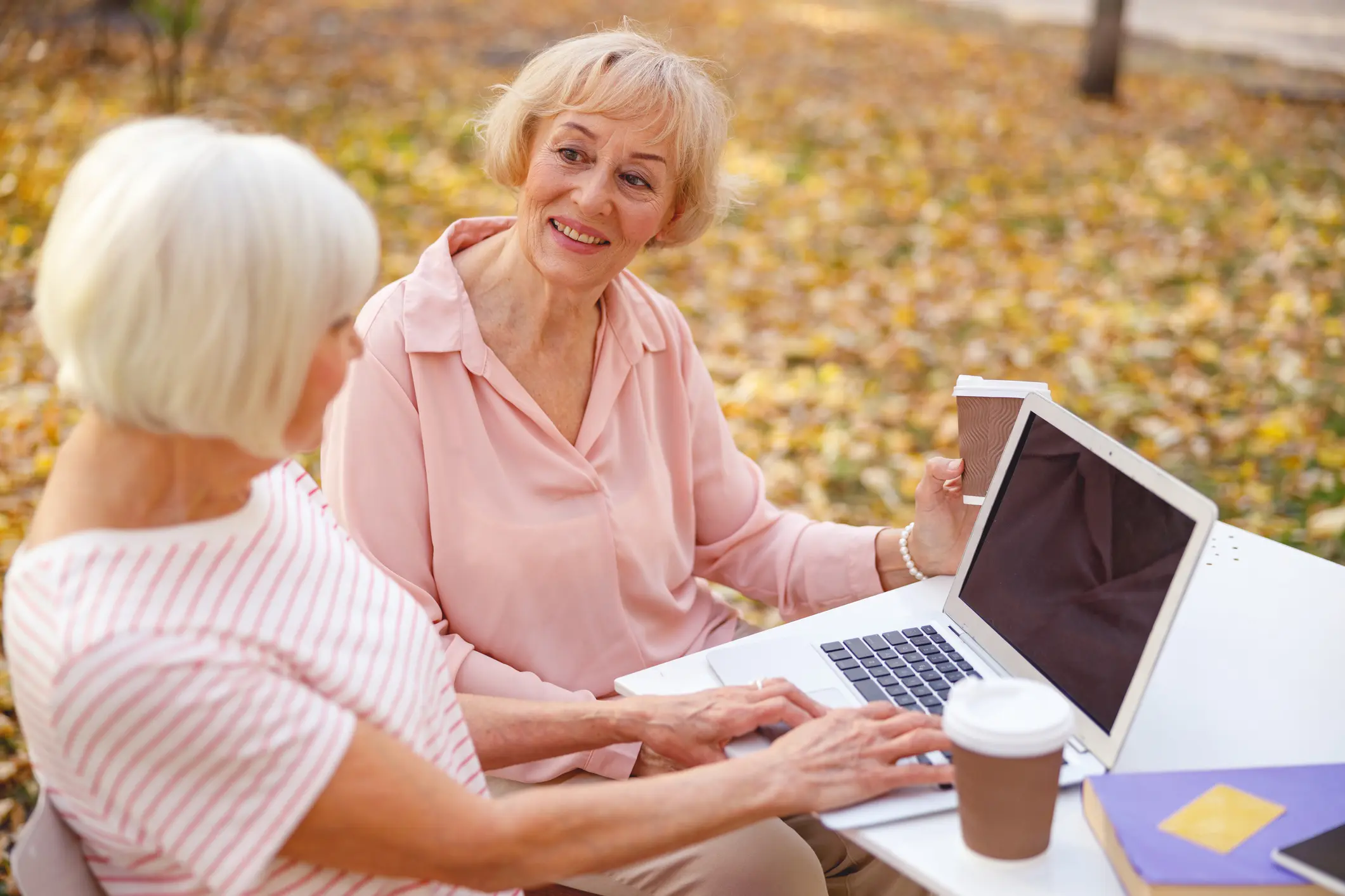 Two elderly women using a laptop outdoors in autumn.