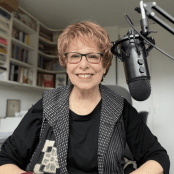 Smiling person with glasses speaking into a microphone in a cozy room.