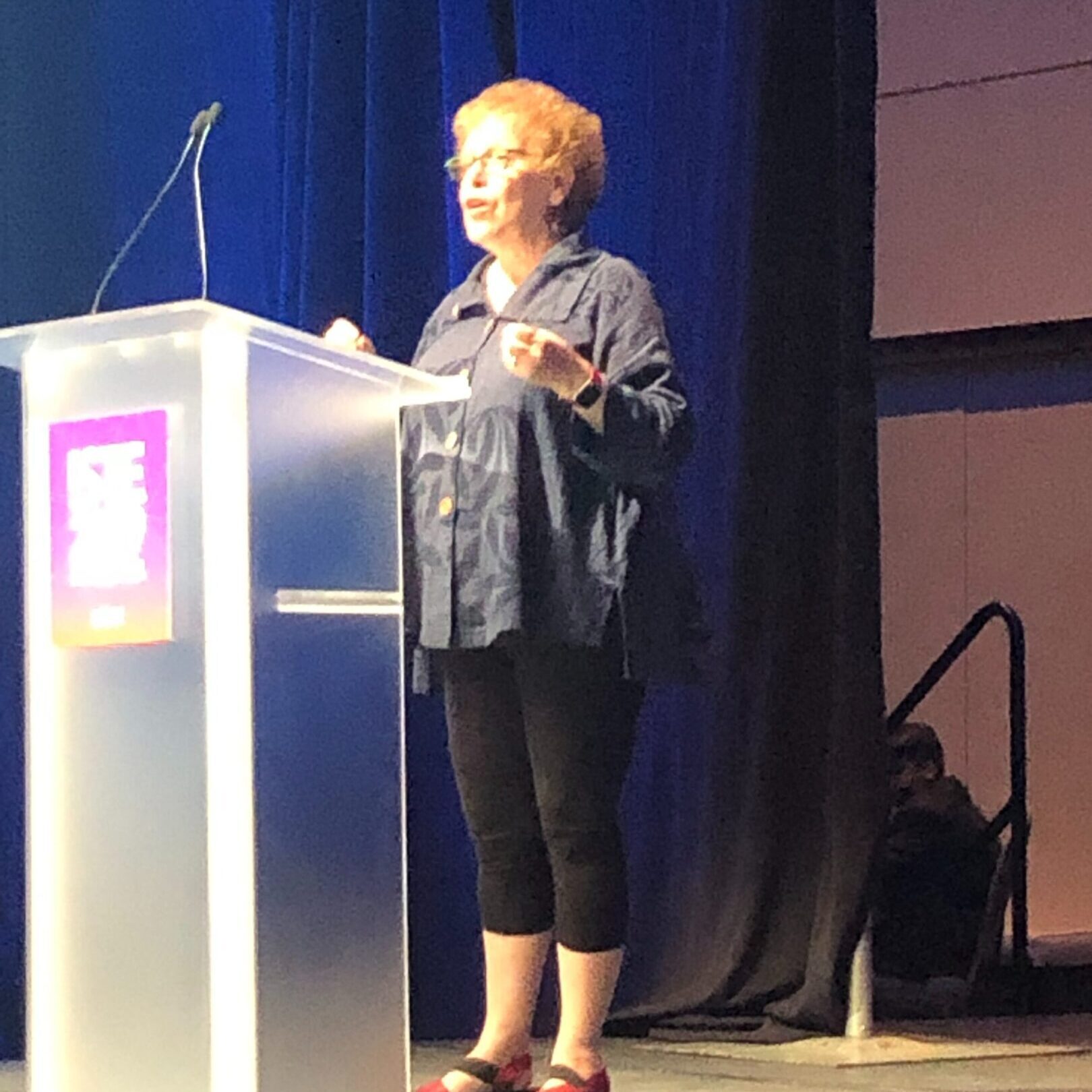 A woman speaks at a podium on stage under bright lighting.