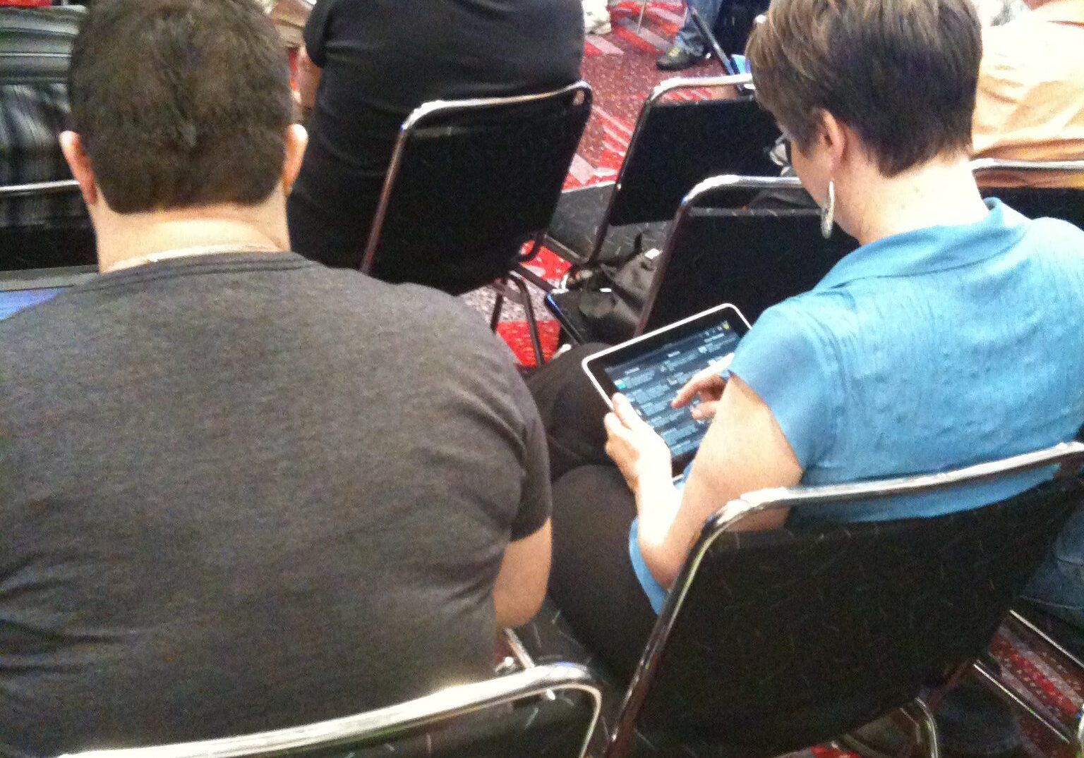 Two men seated, one using a tablet, in a crowded room.